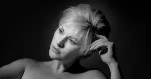 Black and white portrait of a person with short, light-colored hair looking thoughtfully into the distance. They have a neutral expression and are positioned against a dark background, with soft lighting highlighting their face