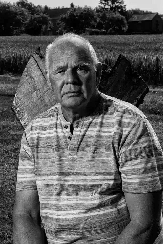 A black and white photo captures Geraint Williams, an elderly man with short hair, sitting in front of a large piece of wood outdoors. He wears a striped shirt and has a serious expression, with a field and farmhouse visible in the background.