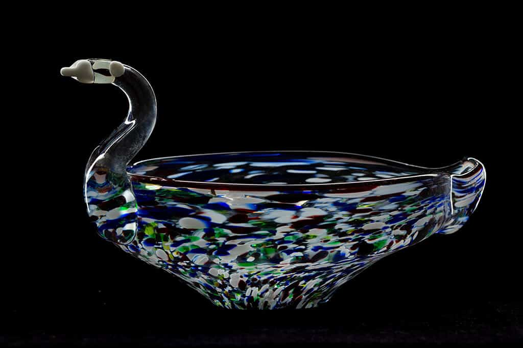 A glass swan-shaped bowl with a long neck and curved body, perfect for product photography. The bowl boasts a multicolored speckled design in shades of blue, green, and white, elegantly set against a dark background.