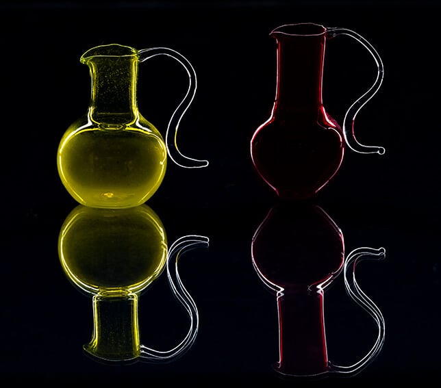 In this stunning example of product photography, two glass pitchers with elegant curved handles are showcased against a black background. One pitcher glows yellow, the other red, each casting a mirrored reflection beneath them.