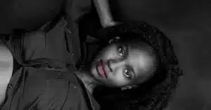 A woman with natural curly hair lies on her back, gazing at the camera. She wears a dark button-up shirt. The black and white photograph emphasizes her striking features and serene expression.