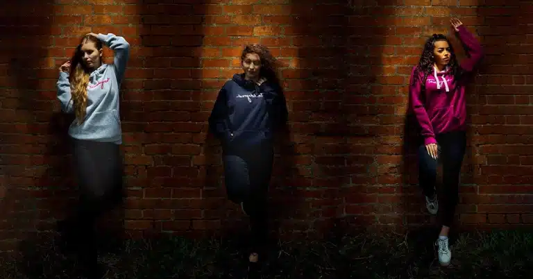 Product Photography Services of three people pose against a brick wall, each illuminated by a spotlight. They wear hoodies in light gray, navy, and magenta. Below them is the word "Teddyred"