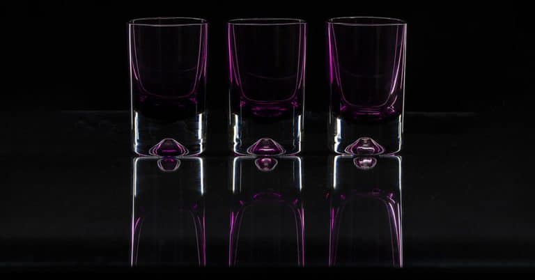 Three empty, transparent shot glasses stand side by side on a reflective surface. A subtle purple hue is visible within each glass, creating an elegant and symmetrical appearance against a dark background.
