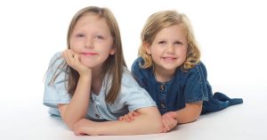 Two young girls with light hair lying on the floor, smiling at the camera. The girl on the left wears a light blue shirt and rests her head on her hand; the girl on the right wears a darker blue dress. The background is white.