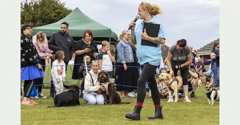 A woman in a light blue shirt speaks into a microphone at an outdoor event. She holds a clipboard and wears black boots. Several people and dogs are gathered around, with some sitting and others standing. A green tent is visible in the background.