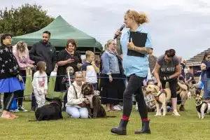 A woman with a microphone walks on grass at an outdoor event with dogs and people gathered around, some seated and some standing near a green marquee.