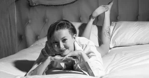 A person with short hair lies on their stomach on a bed, smiling at the camera, with their hands under their chin and their legs bent upward. The image is in black and white.