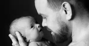 A man smiles gently, holding a baby close. The baby looks up at him, and their faces almost touch. The image is in black and white, highlighting the tender bond between them.