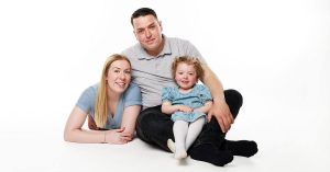 A family of three poses against a white background. An adult woman lies on the floor beside an adult man who sits with a young girl in a blue dress on his lap. All three are smiling at the camera.