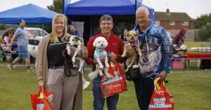 Three smiling adults stand outdoors holding small dogs and red goody bags, with blue tents and people in the background at a community event on a grassy field.