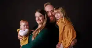 A family of four poses in front of a dark background for a Fourwinds Photography session. The parents, dressed in green and black, each hold a smiling daughter in yellow as they all look warmly at the camera.