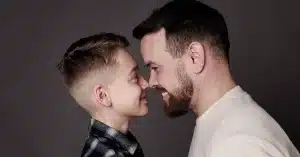 A smiling man and a boy stand face-to-face, touching foreheads and noses against a plain dark background. Capturing their joyful connection, this tender moment highlights the warmth of Northampton Photography.