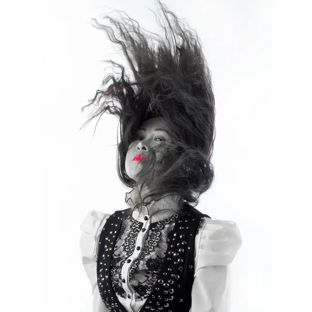 A woman with long dark hair flipping her head upward, her hair suspended in the air. She wears a studded vest over a lace blouse and has bright red lipstick, with a white background behind her.