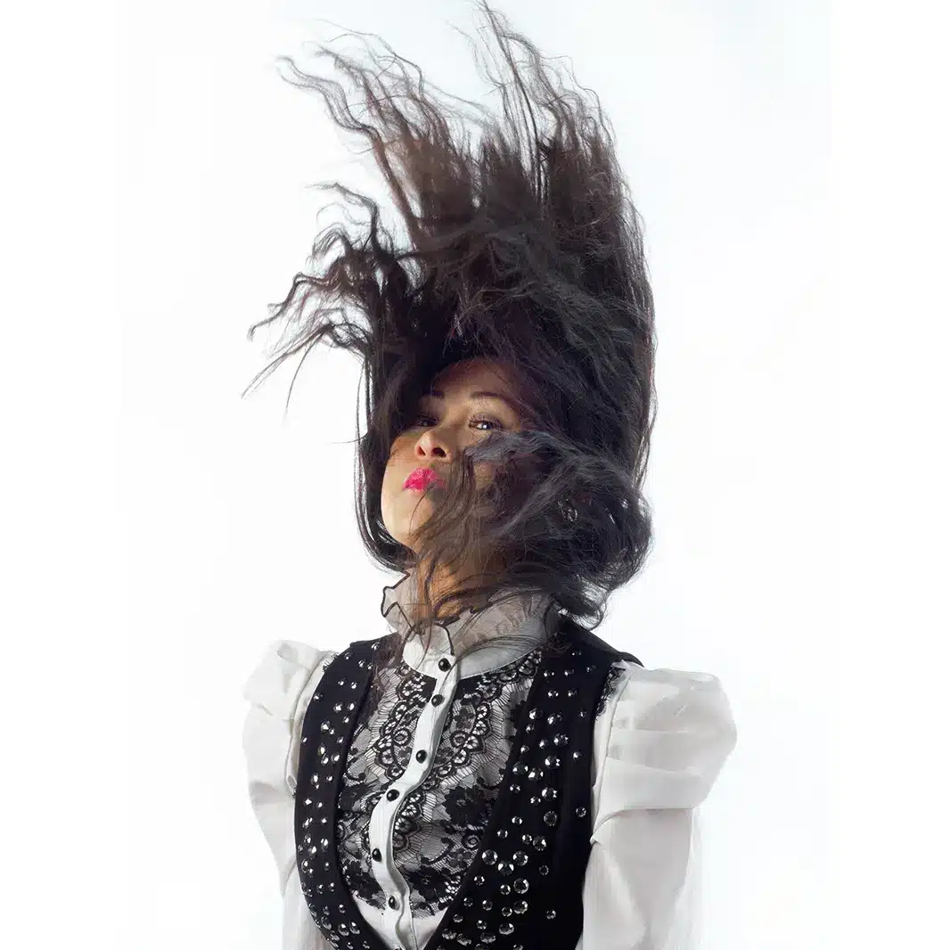 A woman with long dark hair flips her head back, causing her hair to fly upward. She wears a white blouse with lace detail and a black vest decorated with studs. The background is plain white.