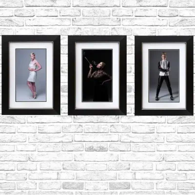 Three framed fashion portraits hang on a white brick wall; the left shows a person in a light dress, the center features a dramatic pose with a cane, and the right displays a person in a suit with hands in pockets. Three framed fashion portraits hang on a white brick wall; the left shows a person in a light dress, the center features a dramatic pose with a cane, and the right displays a person in a suit with hands in pockets.