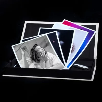 A black box holds several large printed photographs, including a black-and-white portrait of a smiling person talking on the phone. A USB flash drive lies beside the photos, all set against a dark background. A black box holds several large printed photographs, including a black-and-white portrait of a smiling person talking on the phone. A USB flash drive lies beside the photos, all set against a dark background.
