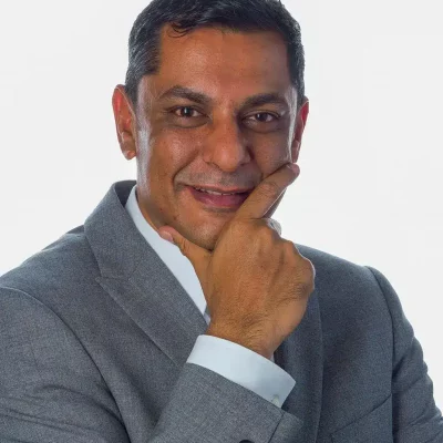 A man in a gray suit smiles, with his hand resting thoughtfully on his chin against a plain white background.