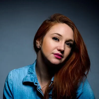 A woman with long red hair and wearing a blue shirt gazes slightly off camera. The background is a smooth gradient of dark grey to light grey. Her expression is calm and confident.