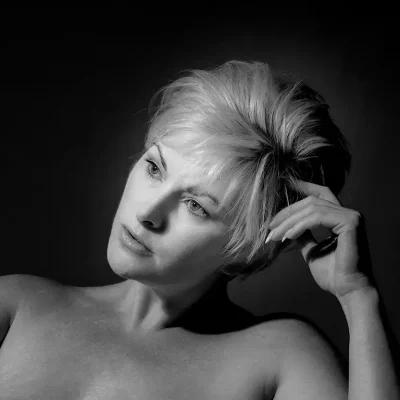 Black and white portrait by Northampton Photography of a light-skinned woman with short, tousled blonde hair, gazing thoughtfully to the side, her hand resting near her head against a dark background. Black and white portrait by Northampton Photography of a light-skinned woman with short, tousled blonde hair, gazing thoughtfully to the side, her hand resting near her head against a dark background.