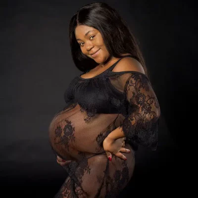 A pregnant woman in a sheer, black lace dress poses against a dark background, smiling with one hand on her hip and the other resting on her belly. A pregnant woman in a sheer, black lace dress poses against a dark background, smiling with one hand on her hip and the other resting on her belly.