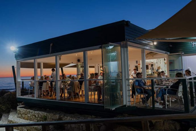 A group of people are dining in a modern, glass-walled restaurant overlooking the ocean at sunset. As an event photographer captures the moment, the sky transitions from deep blue to warm orange hues, and the interior is softly illuminated, creating a cozy atmosphere.