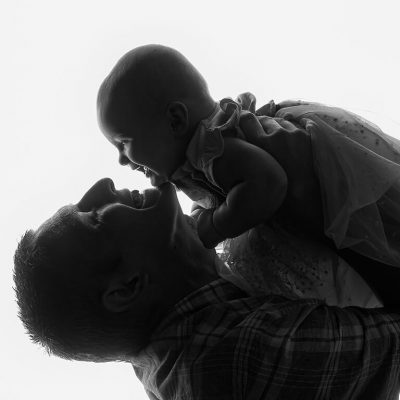 A black and white silhouette of an adult holding a smiling baby up in the air. Both appear joyful, with the baby reaching toward the adult’s face and the adult looking up at the baby. A black and white silhouette of an adult holding a smiling baby up in the air. Both appear joyful, with the baby reaching toward the adult’s face and the adult looking up at the baby.