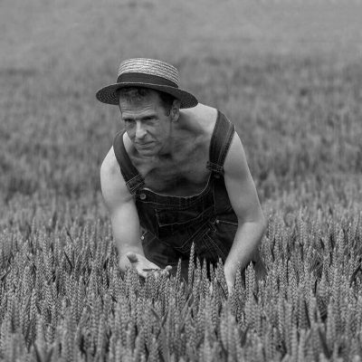 Black and white image of a weathered farmer wearing overalls and a straw hat, inspecting a field of wheat. He is kneeling, surrounded by crop rows, examining the plants closely. For more on this hardworking spirit or related inquiries, feel free to contact us.