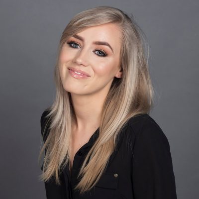A young woman with long, straight blonde hair and light makeup smiles slightly while wearing a black button-up shirt, posing in front of a plain gray background. A young woman with long, straight blonde hair and light makeup smiles slightly while wearing a black button-up shirt, posing in front of a plain gray background.