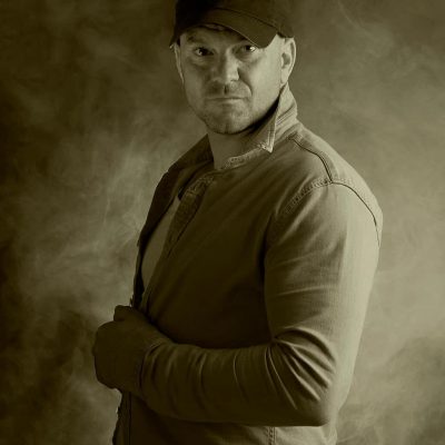 A man wearing a baseball cap and a button-up shirt stands against a smoky, sepia-toned background, looking seriously at the camera with one hand holding his shirt collar. A man wearing a baseball cap and a button-up shirt stands against a smoky, sepia-toned background, looking seriously at the camera with one hand holding his shirt collar.
