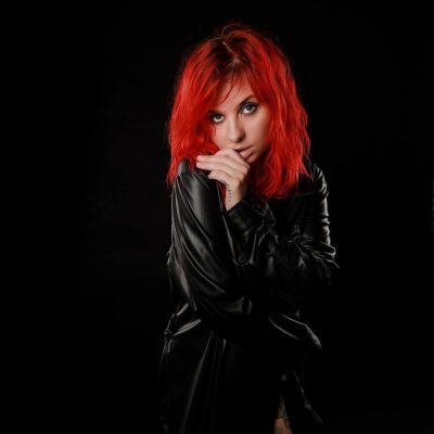 Portfolio Photography of a Woman with vibrant red hair stands against a dark background, wearing a black leather jacket.