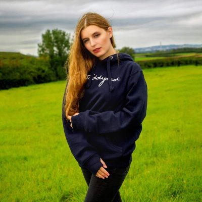 Product Photography Service of A person with long brown hair stands in a grassy field, wearing a dark hoodie adorned with cursive writing
