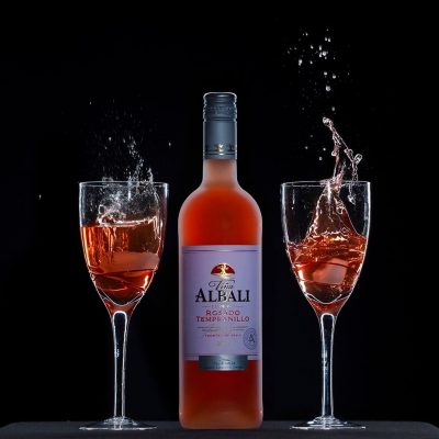 A bottle of Albali Rosado Tempranillo wine is centered between two glasses filled with the same rosé, capturing an exquisite moment in product photography. The splashes of liquid against the black background suggest movement and energy.