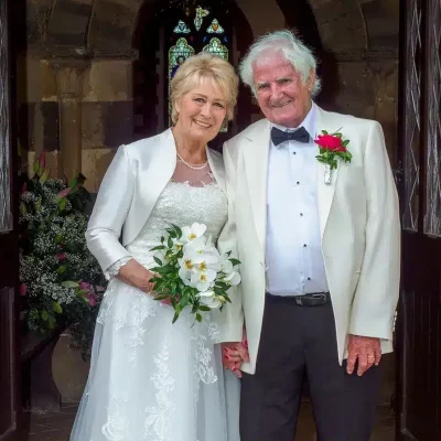 An elderly couple stands in a church doorway, smiling. The woman wears a white wedding dress and holds a bouquet, while the man is dressed in a white jacket, black pants, and a bow tie. They appear to be newlyweds. An elderly couple stands in a church doorway, smiling. The woman wears a white wedding dress and holds a bouquet, while the man is dressed in a white jacket, black pants, and a bow tie. They appear to be newlyweds.