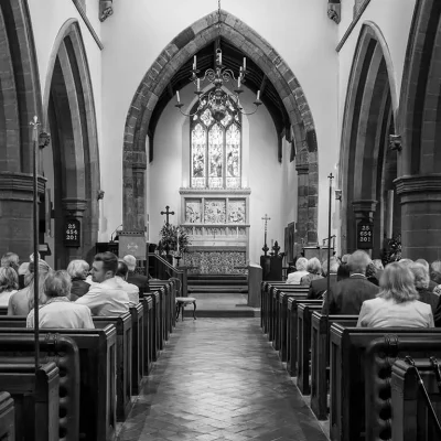 Black and white photo of people seated in pews inside a church, facing the altar at the front, with arched doorways and stained glass windows visible. Black and white photo of people seated in pews inside a church, facing the altar at the front, with arched doorways and stained glass windows visible.